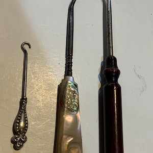 May include: Three vintage crochet hooks. The hooks are made of metal and have decorative handles. One hook has a small, ornate silver handle. Another has a mother-of-pearl handle with abalone inlay. The third has a dark wood handle.