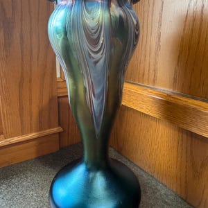 Op de afbeelding: Een iriserende glazen vaas met een uitlopende, gerimpelde rand. De vaas loopt over van een diepe teal basis naar een wervelend patroon van groen, blauw en zilver. De vaas heeft een bolvormige basis en een slanke hals.