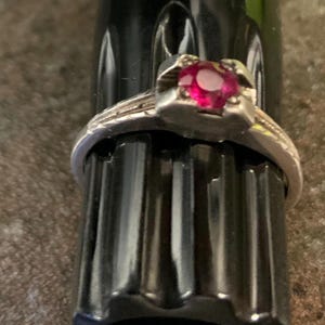 May include: A silver ring with a vibrant red gemstone at its center. The ring features a detailed band with delicate patterns. The gemstone is set in a square, pronged setting, showcasing its color and brilliance. The ring is a piece of jewelry.