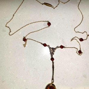May include: A vintage gold-toned necklace with a pendant locket. The necklace features red gemstone accents and a decorative filigree design. The locket is oval-shaped with an amber-colored center.