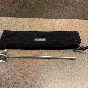 May include: A silver candle snuffer with a bell-shaped head and a long handle, alongside a black velvet pouch. The pouch features a small rectangular label with text. The snuffer is designed for safe candle extinguishing.