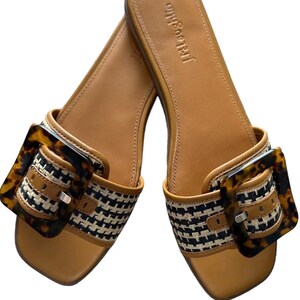 May include: Tan leather sandals with a woven black and white strap and a tortoiseshell buckle. The sandals have a square toe and the brand name "J.McLaughlin" is printed on the footbed.