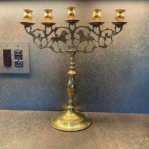 May include: A brass menorah with six candle holders on top and a central holder. The base is round and the stem is ornate. The menorah is on a countertop with a textured surface.