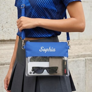May include: A clear crossbody bag with a blue top section and strap. The bag is filled with items including a phone, sunglasses, and a wallet. The name "Sophia" is embroidered in white on the blue section. The bag is worn over the shoulder.