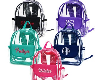 May include: Five clear backpacks with various colored accents. Each backpack has a front pocket with a name or initials embroidered on it. The backpacks are in black, purple, navy, teal, and pink.