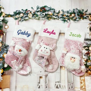 May include: Three pink velvet Christmas stockings with white fur trim. The stockings are personalized with the names "Gabby", "Layla", and "Jacob". Each stocking features a different festive character: a snowman, a reindeer, and Santa Claus.