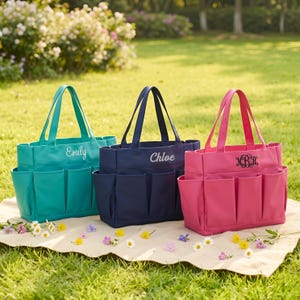 May include: Three tote bags in teal, navy, and pink, each with multiple exterior pockets. The teal bag has "Emily" embroidered, the navy bag has "Chloe", and the pink bag has a monogram. The bags are on a beige blanket on a grassy lawn.