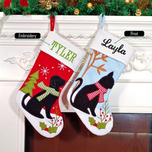 May include: Two personalized Christmas stockings with embroidered names "Tyler" and "Layla". The stocking on the left is red with a black dog and a green tree. The stocking on the right is blue with a black cat and a white tree. The stockings have white cuffs with the names embroidered in green.