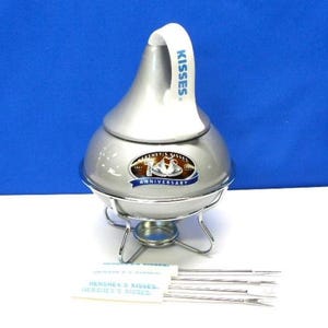 May include: A silver Hershey's Kisses fondue pot with a white handle that reads "Kisses." The pot has a silver base and a metal stand. The pot has a Hershey's Kisses Anniversary logo. Includes skewers.