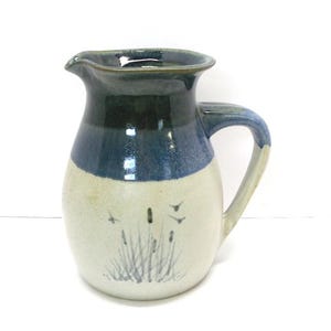 May include: A ceramic pitcher with a dark blue rim, a blue band, and a cream-colored base. The base features a hand-painted design of reeds and birds. The pitcher has a curved handle and a spout for pouring.