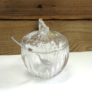 May include: A clear glass sugar bowl with a lid and a small spoon. The bowl has a decorative, faceted design.