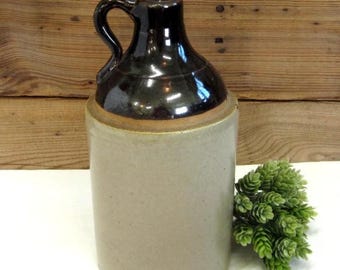 May include: A vintage stoneware jug with a dark brown glazed top and handle, and a light gray body. The jug has a classic, rustic design, perfect for farmhouse decor. A small green plant is next to the jug.