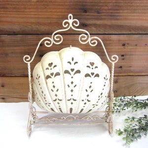 May include: A cream-colored metal shell-shaped basket suspended within a decorative arched frame. The shell features intricate cut-out patterns including small heart shapes and vertical slits. The metal frame has scrolled details and a distressed finish.