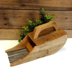 May include: A rustic wooden berry picker with a curved design and a handle on top. The tool features metal tines for harvesting berries and is made of weathered wood, showcasing natural grain patterns. The background is a wooden surface.