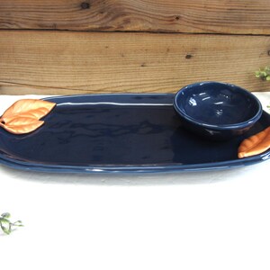 May include: A navy blue ceramic serving platter with a small matching bowl. The platter has a long oval shape with a copper-colored leaf accent on each end. The bowl sits on the platter, creating a cohesive serving set.