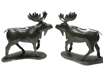 May include: Two metal moose sculptures facing each other. The moose are made of metal and have a dark gray finish. They are standing on a metal base.