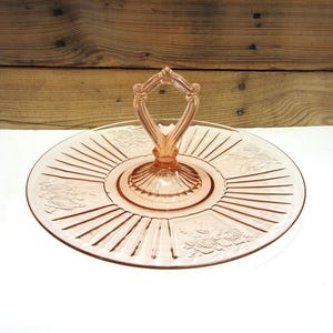 May include: A vintage pink glass serving tray with a central handle. The tray features a circular design with radial lines and floral patterns. The handle has a decorative, openwork design.