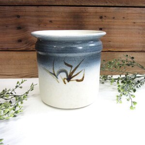 May include: A ceramic stoneware crock with a white base and a blue-gray top. The crock features a hand-painted brown floral design. The rustic kitchenware is suitable for storing utensils or as a decorative piece.