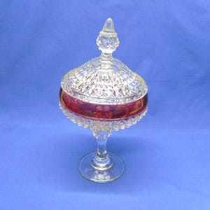 May include: A clear glass candy dish with a lid, featuring a ruby red band around the bowl. The dish has a pedestal base and a diamond-cut pattern on the lid and bowl. The finial on the lid is pointed.
