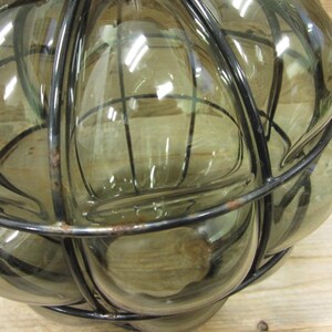 Puede incluir: Una botella de vidrio decorativa con un marco de metal en forma de jaula. La botella est&aacute; hecha de vidrio transparente de color verde oliva y tiene una forma redondeada. El marco de metal es negro y rodea el vidrio, creando un dise&ntilde;o &uacute;nico. La botella probablemente se utiliza con fines decorativos.