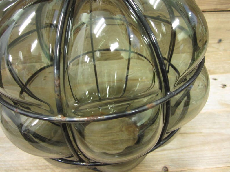 Puede incluir: Una botella de vidrio decorativa con un marco de metal en forma de jaula. La botella est&aacute; hecha de vidrio transparente de color verde oliva y tiene una forma redondeada. El marco de metal es negro y rodea el vidrio, creando un dise&ntilde;o &uacute;nico. La botella probablemente se utiliza con fines decorativos.