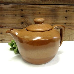 May include: A brown ceramic teapot with a lid and handle. The teapot has a glossy finish and a rounded shape. The background is a wooden surface.