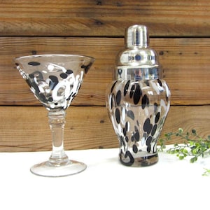 May include: A clear cocktail shaker and matching martini glass, both adorned with a black and white spotted pattern. The shaker has a silver lid, and the glass has a long stem. The set is displayed on a white surface.
