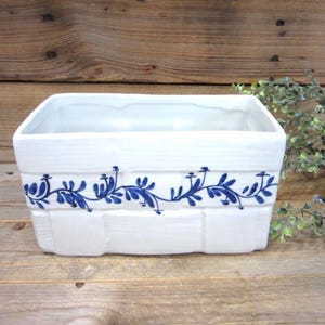 May include: A rectangular white ceramic planter with a basket-weave texture on the exterior. The planter features a hand-painted blue floral vine pattern encircling the middle section. The interior is smooth, white, and hollow with an open top.