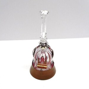 May include: A clear crystal bell with a faceted handle and a ruby-colored interior. The bell has a wooden base and a small rectangular label. The bell is approximately 6 inches tall.