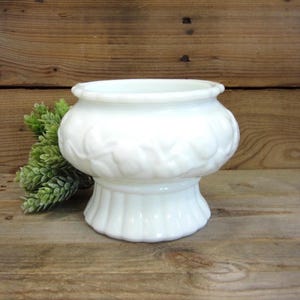 May include: A white milk glass planter with a decorative leaf pattern around the bowl and a ribbed base. The planter is set against a wooden background with a sprig of green foliage.