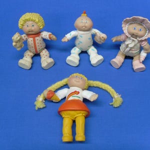 May include: Four Cabbage Patch Kids dolls on a blue background. The dolls have various outfits and hairstyles. One doll holds a teddy bear. The dolls are made of plastic and have painted faces. The dolls are vintage and collectible.