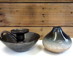 May include: Two ceramic pieces: a dark gray candle holder with a handle and a small, round, dark blue and beige vase. The candle holder has a circular base and a raised center for the candle. The vase has a gradient color scheme.