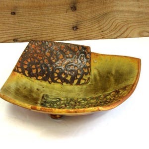 May include: A handcrafted ceramic bowl with a unique design. The bowl features a square section with a textured, brown pattern, and a curved, olive-green body. The bowl is approximately 10 inches long and 8 inches wide.