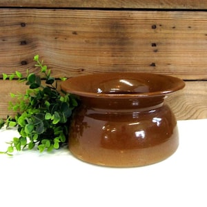 May include: A brown ceramic pot with a wide rim and a rounded body. The pot has a glossy finish and sits on a white surface. Green leafy sprigs are to the left of the pot. The background is a wooden wall.