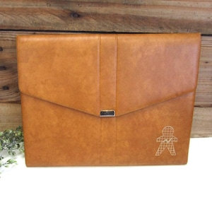 May include: A tan leather portfolio with a flap closure and a silver-tone clasp. The front features a stitched design and a small, embroidered figure. The portfolio is rectangular and appears to be new.
