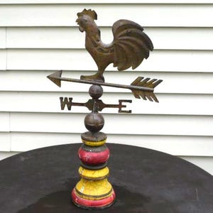 May include: A metal weather vane featuring a rooster silhouette mounted on an arrow with directional markers W and E. The base is painted with alternating yellow and red rings, showing a weathered, cracked finish on the painted surface.