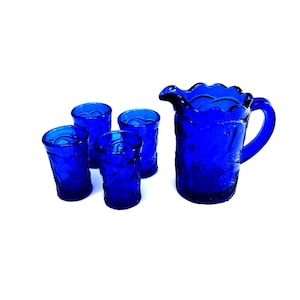 May include: A set of cobalt blue glass pitcher and four tumblers. The pitcher has a scalloped rim and a handle. The tumblers are all the same size and have a textured surface.