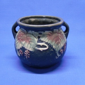 May include: A small, dark blue ceramic pot with two handles. The pot is decorated with a raised design of green leaves and purple berries.
