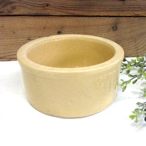 May include: A round, beige stoneware bowl with a wide rim. The bowl has a slightly textured surface and a simple, classic design. The interior is smooth and the bowl is empty. The background is a wooden surface.