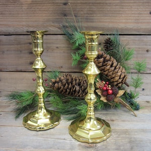May include: A pair of gold-toned brass candle holders with a fluted design. The candle holders are sitting on a wooden surface with pine branches and pine cones.