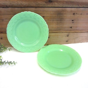 May include: Two jadeite green plates with a floral embossed border. The plates are round with a slightly scalloped edge. The background is a wooden surface.