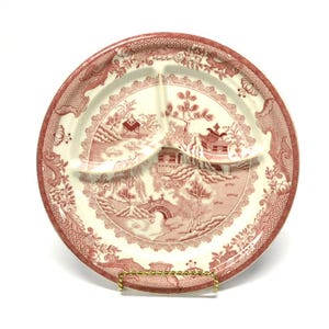 May include: A round, divided plate with a red and cream-colored design. The plate features a landscape scene with buildings, trees, and a bridge. The rim has a decorative border. The plate is on a gold-colored metal stand.