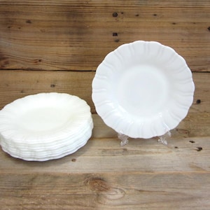 May include: A stack of white ceramic dinner plates with scalloped edges. The plates are stacked on a wooden surface.