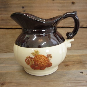 May include: A brown and white ceramic pitcher with a pineapple and fruit design. The pitcher has a brown handle and a wide mouth.