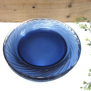 May include: A stack of four cobalt blue glass plates. Each plate has a spiral design around the rim. The plates are on a white surface with a wooden background and green foliage.