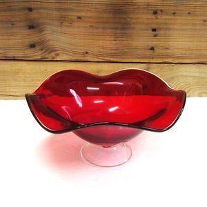 May include: A vintage, ruby red glass bowl with a scalloped rim and a clear, pedestal base. The bowl has a glossy finish and a dark rim. The background is a wooden surface.