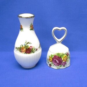 May include: A white porcelain bud vase and a matching bell, both featuring a floral pattern of red, yellow, and pink roses with green leaves. The vase has a ribbed base and gold trim, while the bell has a heart-shaped handle and gold trim.