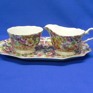 May include: A floral-patterned tea set featuring a teacup, creamer, and a serving tray. The set has a white base with a colorful floral design in pink, yellow, and purple. The edges are trimmed with gold.