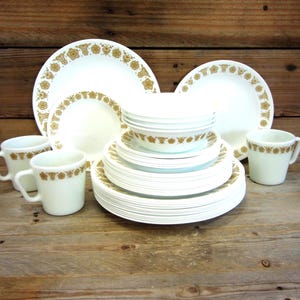 May include: A complete vintage white dinnerware set with a gold floral pattern. The set includes plates, bowls, and mugs. The plates and bowls are stacked, and the mugs are arranged in front. The set is displayed on a wooden surface.
