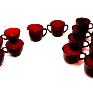 May include: A collection of ruby red glass teacups and saucers, arranged on a white surface. The set includes various cup styles, including some with handles and a creamer. The cups have a glossy finish.